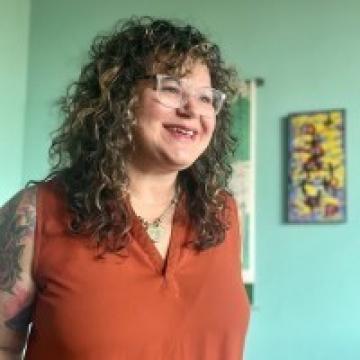 A woman at a side angle with curly brown hair wearing a orange shirt and glasses, smiling against a green backgorund.