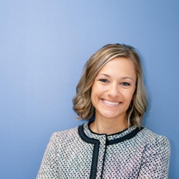 White woman with short dark blonde hair smiling at the camera, standing in front of a blue background wearing a gray jacket with her arms crossed.