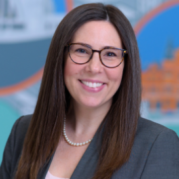 A woman with dark hair, wearing glasses, smiling in front of a blue and orange background.