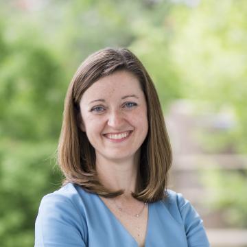 White woman with shoulder length brown hair wearing a blue dress with arms crossed smiling at the camera.